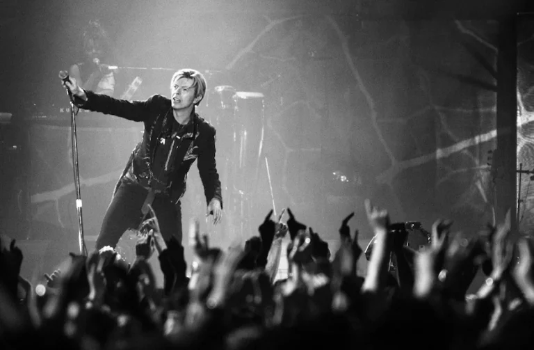 Black and white photo of david bowie leaning toward a cheering crowd, holding a microphone.